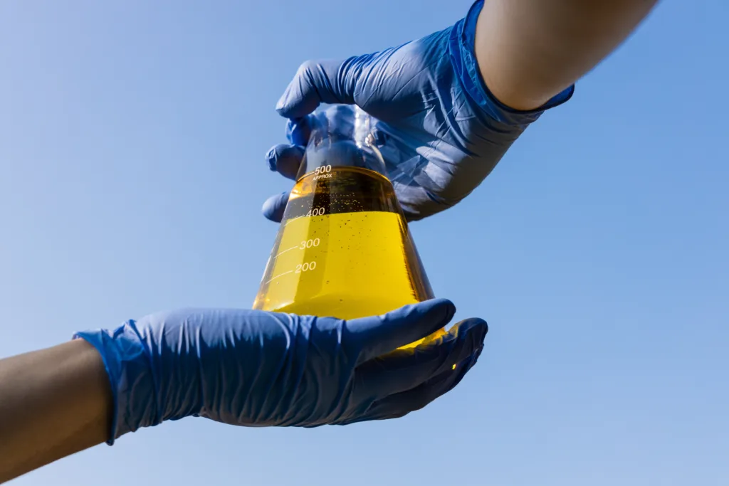 A person wearing blue nitrile gloves is holding a glass chemistry flask up to a blue sky. The flask contains yellow ethanol.